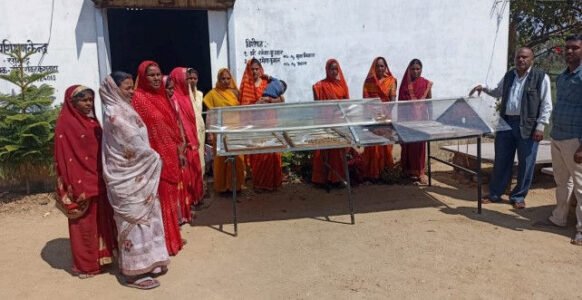 Women in red and orange sarees standing beside a solar dryer during a rural demonstration